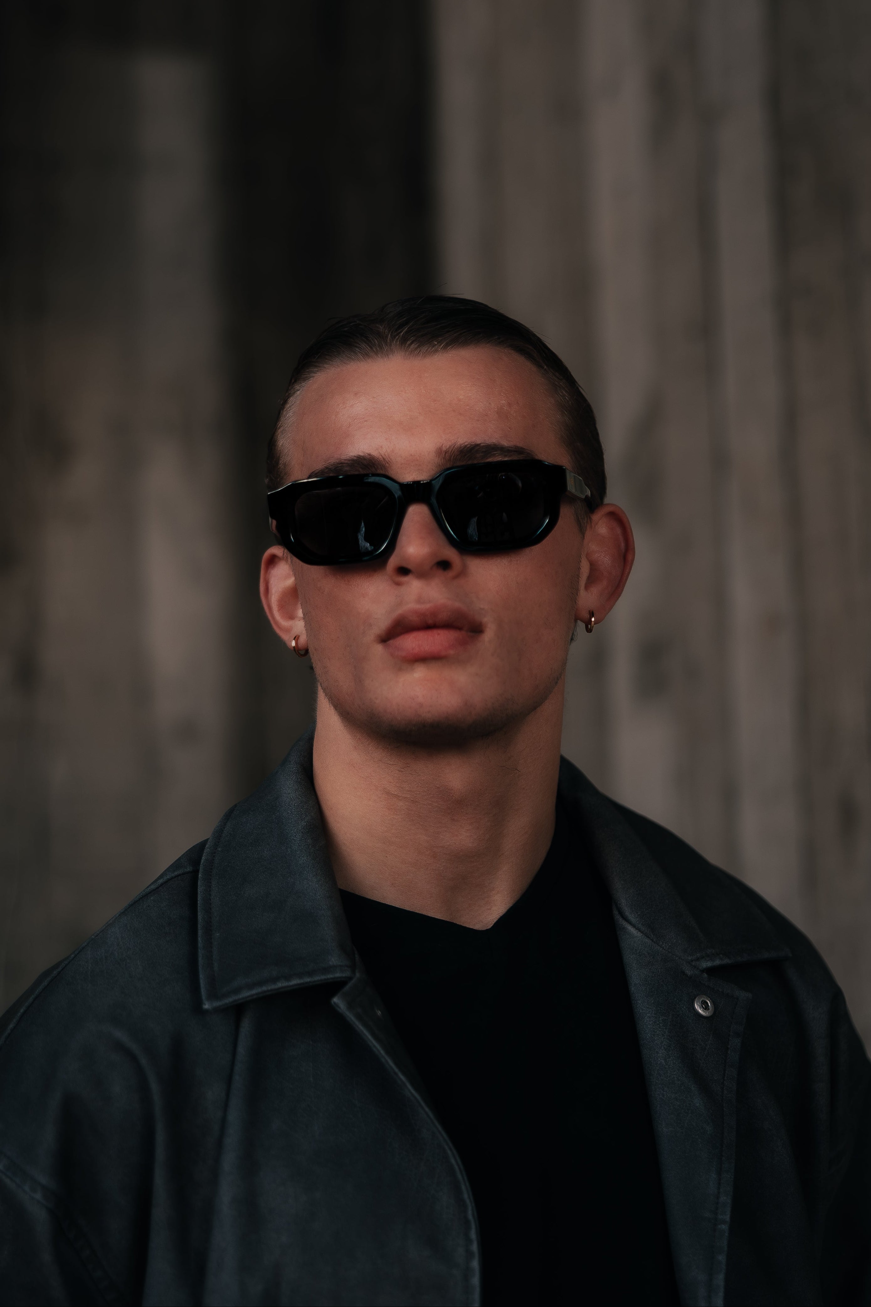 Man wearing dark sunglasses and a dark jacket against a textured wall.