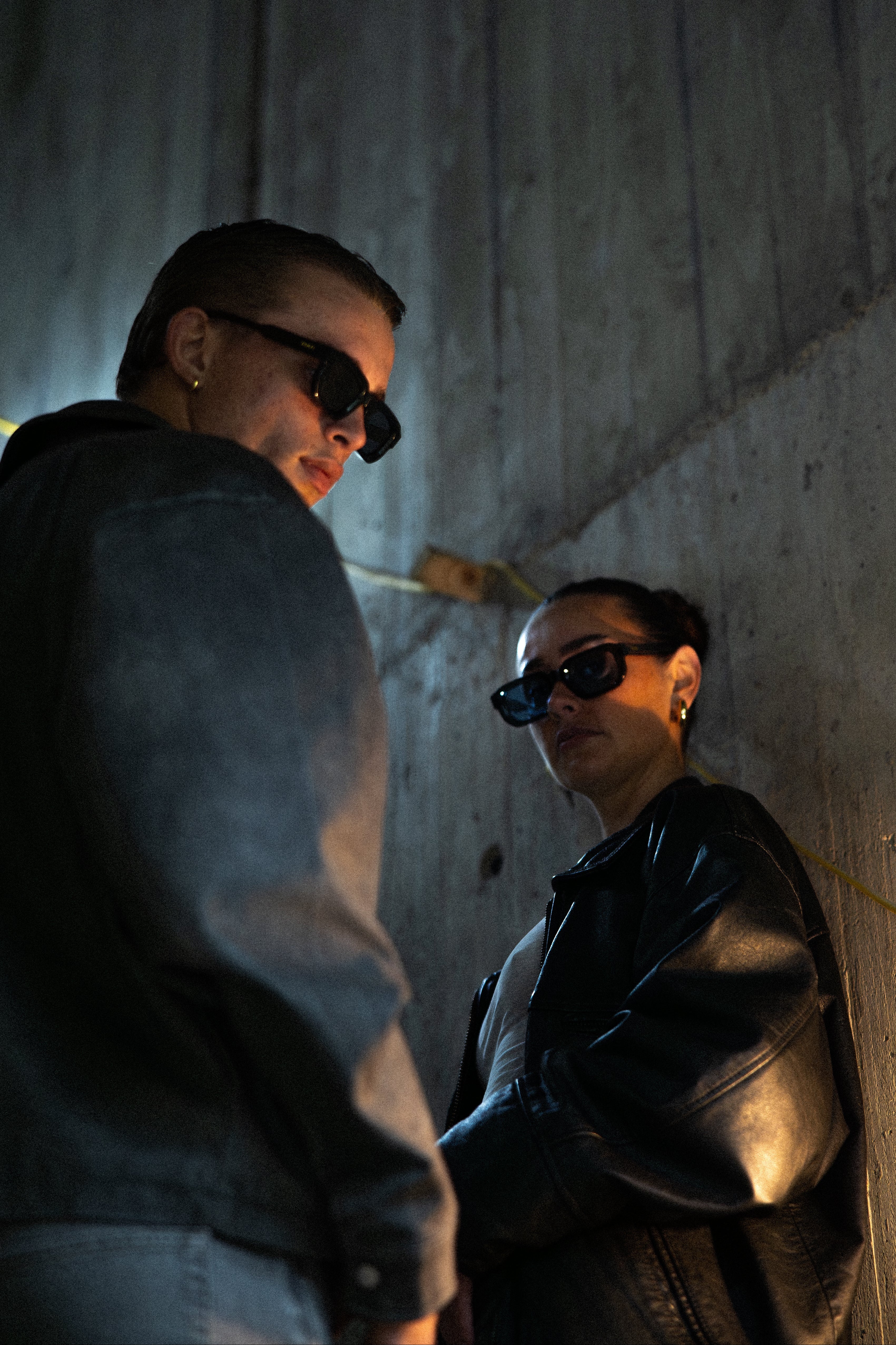 Two people wearing sunglasses in a dimly lit setting with concrete walls.