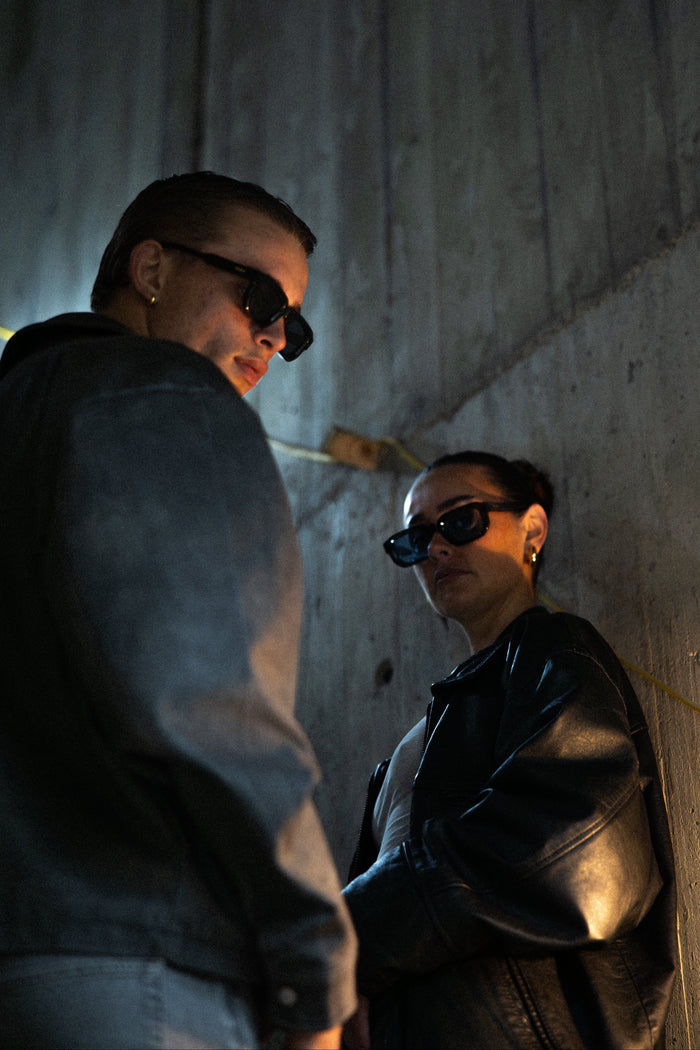 Two people wearing sunglasses in a dimly lit setting with concrete walls.