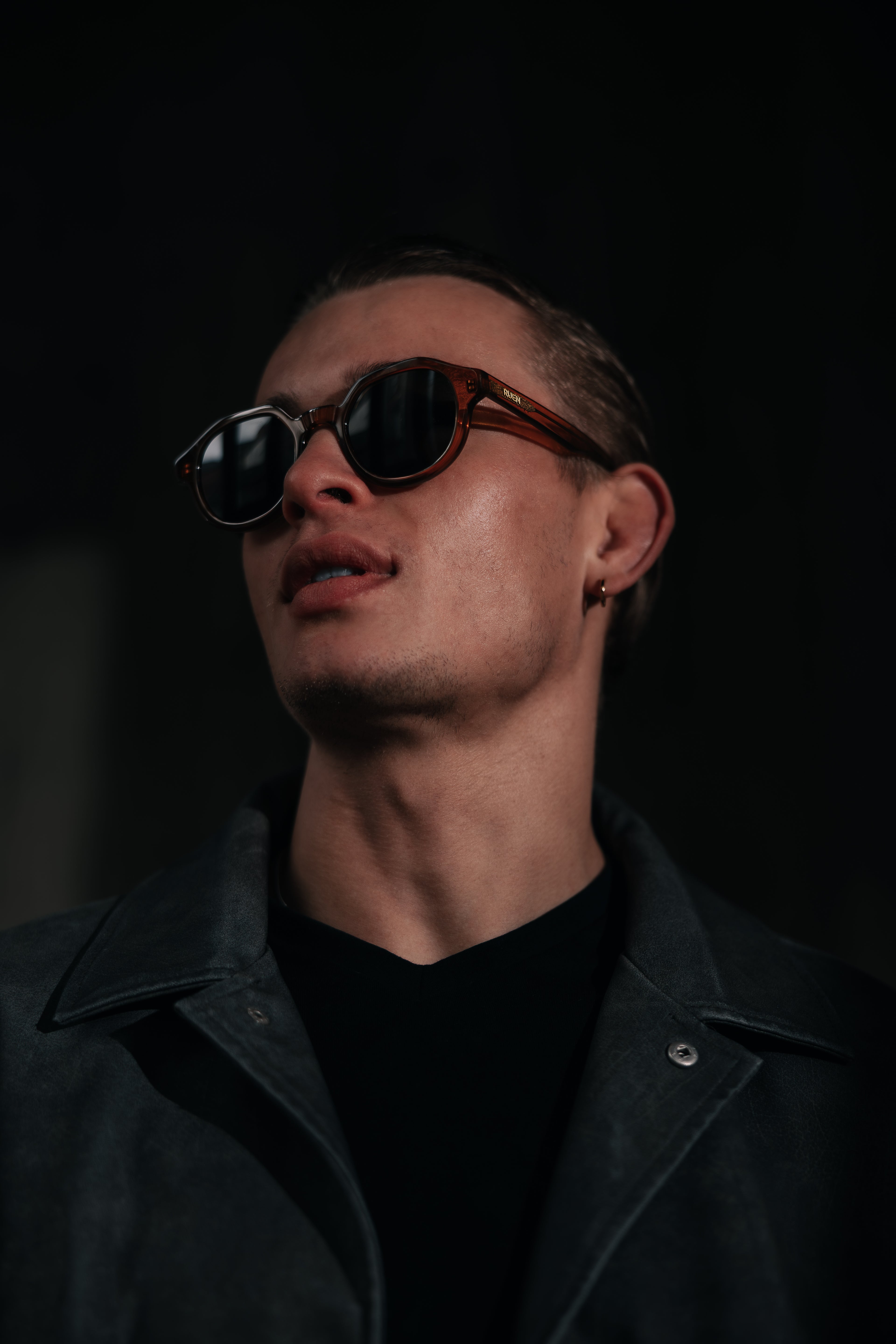 Man wearing sunglasses and a dark coat against a black background