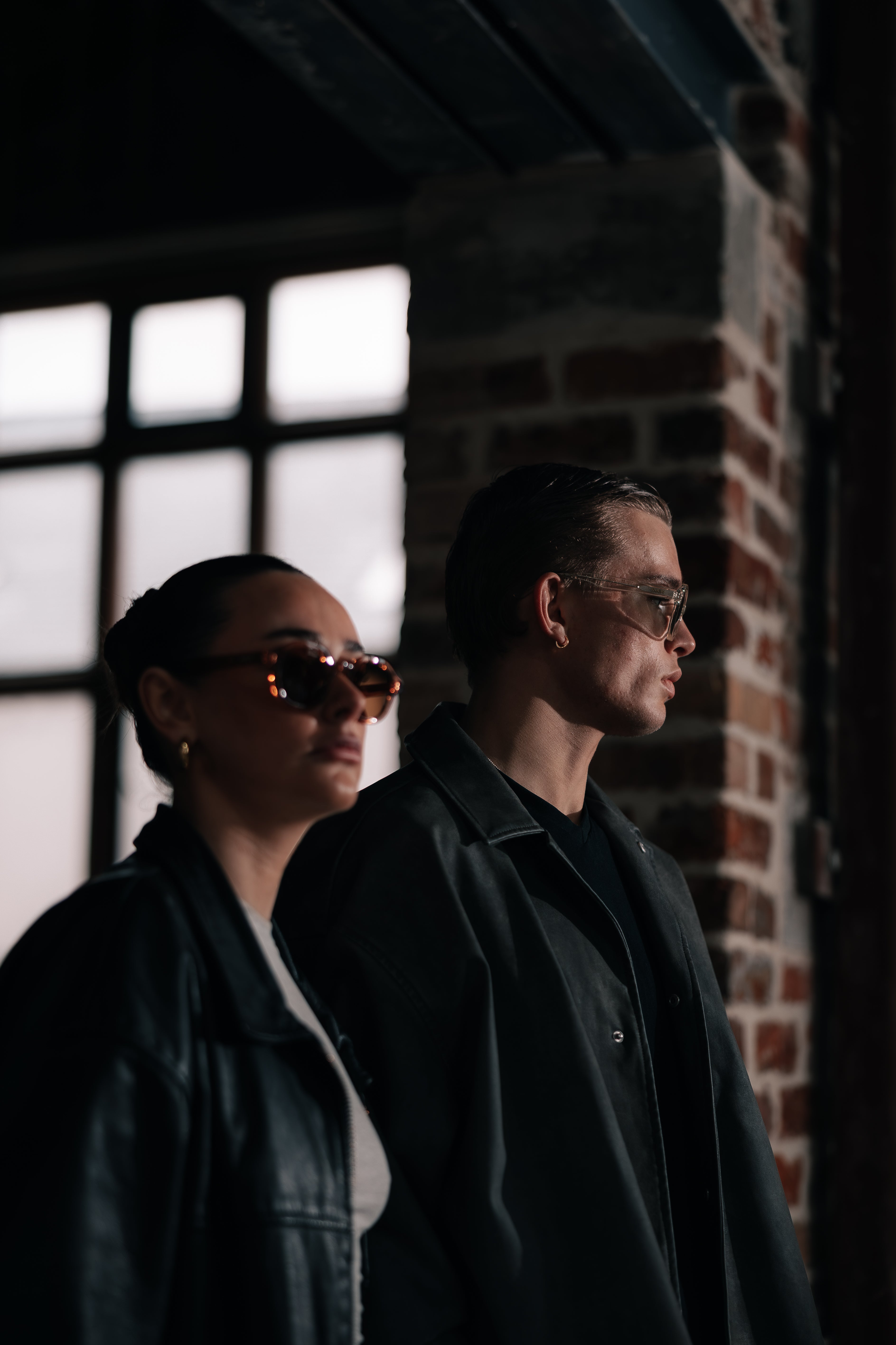 Two people wearing sunglasses standing in front of a brick wall.